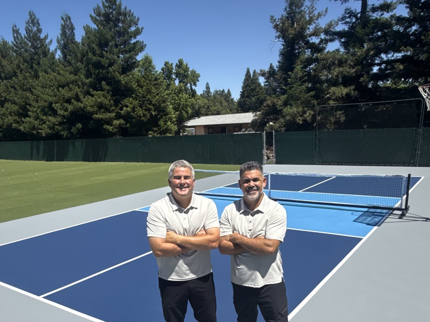 The two owners of NorCal Surfaces, arms crossed, on a blue pickleball court they built